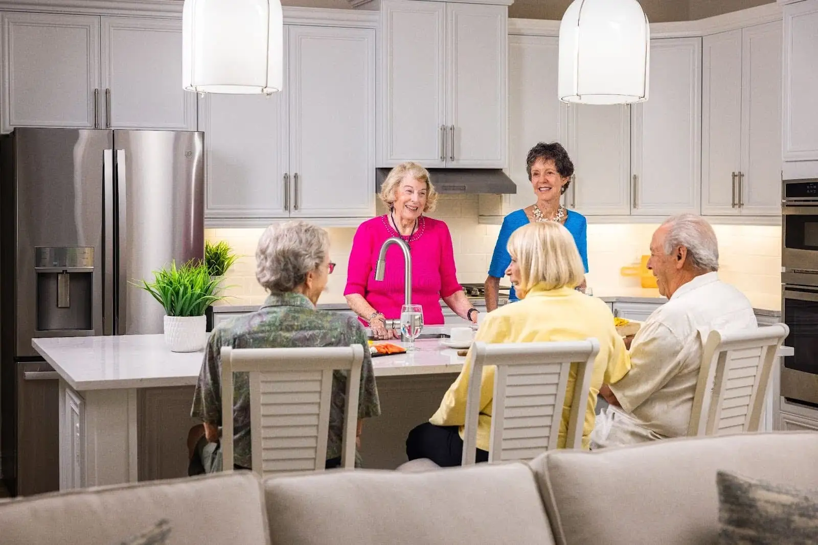 A senior group sitting and standing around a kitchen island, talking about life plan community benefits at The Arlington Of Naples, FL.